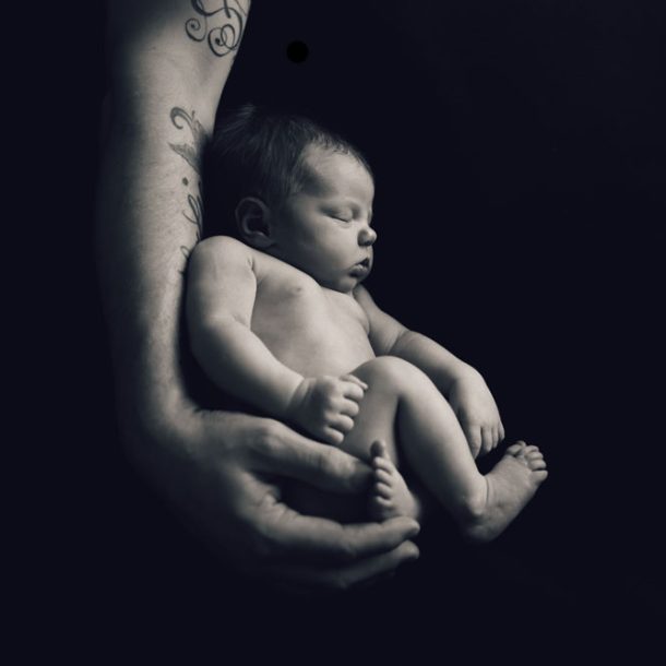 Nyfödd bebis sover tryggt i en förälders tatuerade armar, fångad i en konstnärlig svartvit nyföddfotografering.